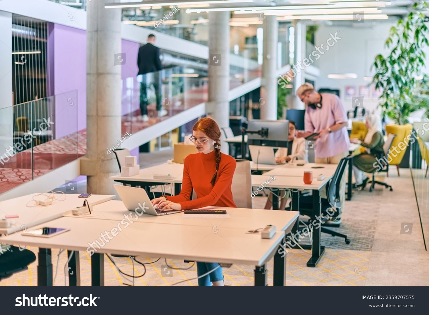 In a modern startup office, a professional businesswoman with orange hair sitting at her laptop, epitomizing innovation and productivity in her contemporary workspace. #2359707575