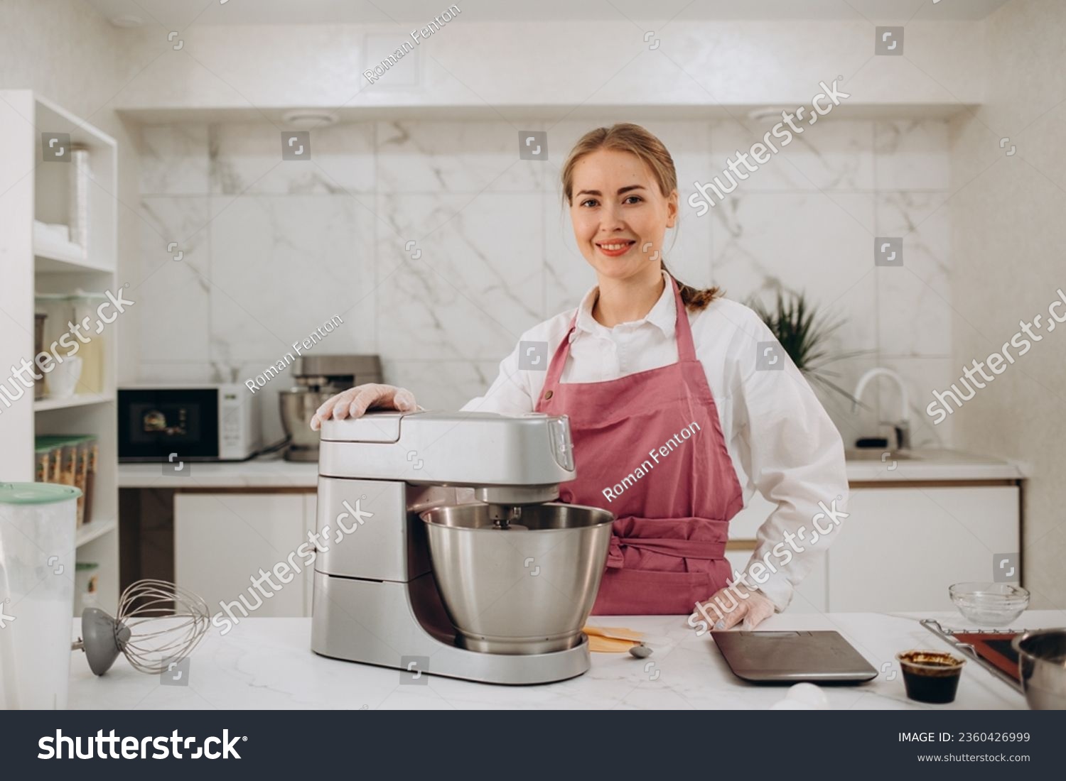 Front view of pretty joyful young woman, professional confectioner in white uniform and hat, using an electric planetary mixer for whipping egg whites, pouring sugar into the bowl.  #2360426999