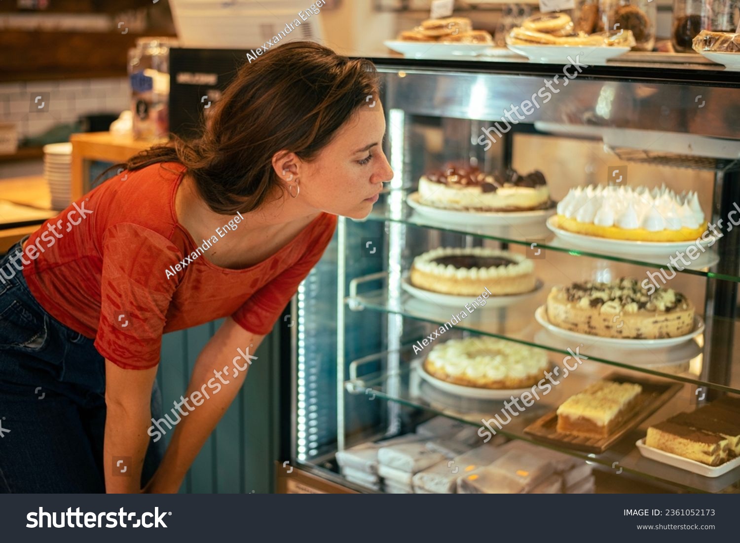 Beautiful 25-year-old woman, white with freckles, wearing jeans and a red t-shirt, in a specialty café, placing her order, choosing a sweet cake to eat. She's in a modern café #2361052173
