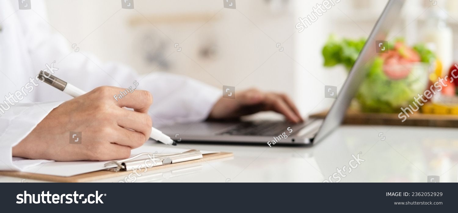 Close-up of dietitian hand writing with laptop giving online weight loss and healthy diet consultation with fresh vegetable fruit measuring tape and glass of clean water on desk against #2362052929