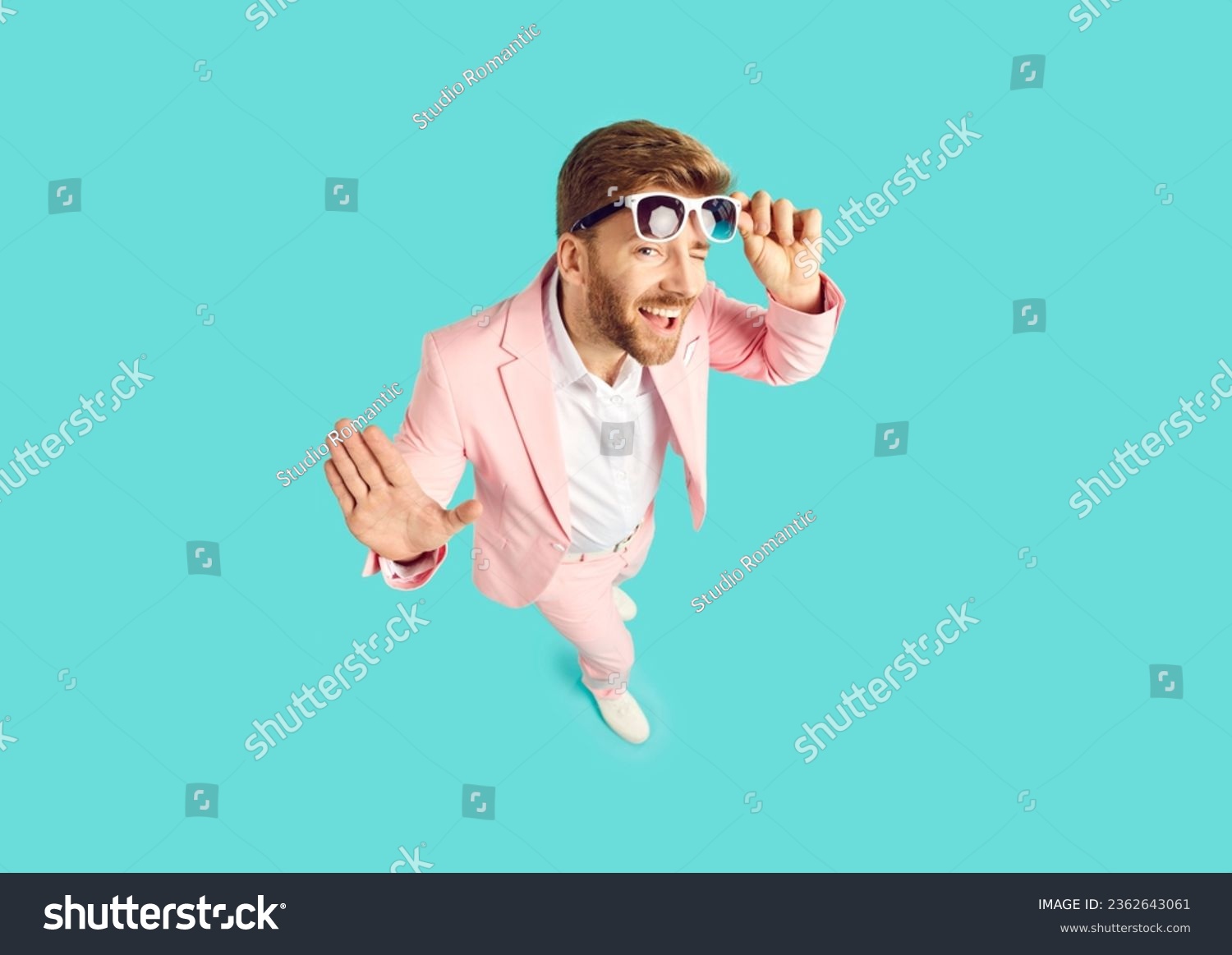 High angle shot from above of happy cheeky handsome young man wearing pink suit standing on blue color background looking at camera, smiling, taking off sunglasses and winking his eye. Fashion concept #2362643061