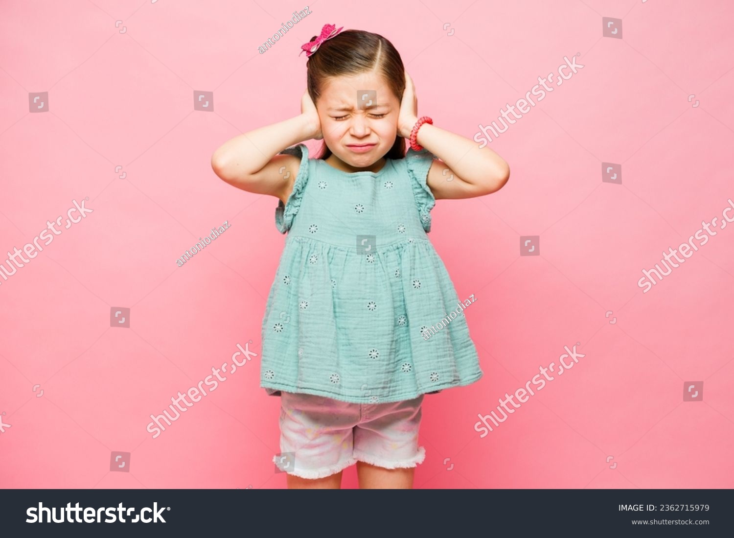Annoyed elementary little kid feeling upset because of loud noises covering her ears against a pink studio background #2362715979