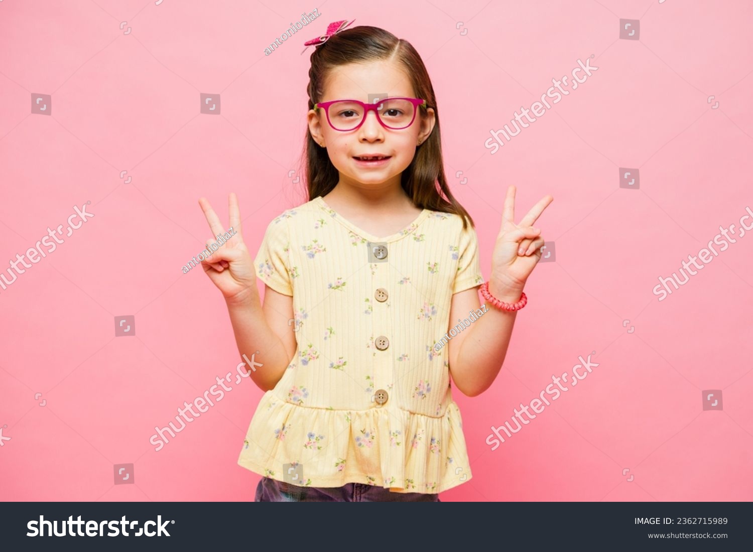 Cheerful little girl making the peace sign and having fun playing while smiling looking happy in a pink studio background #2362715989