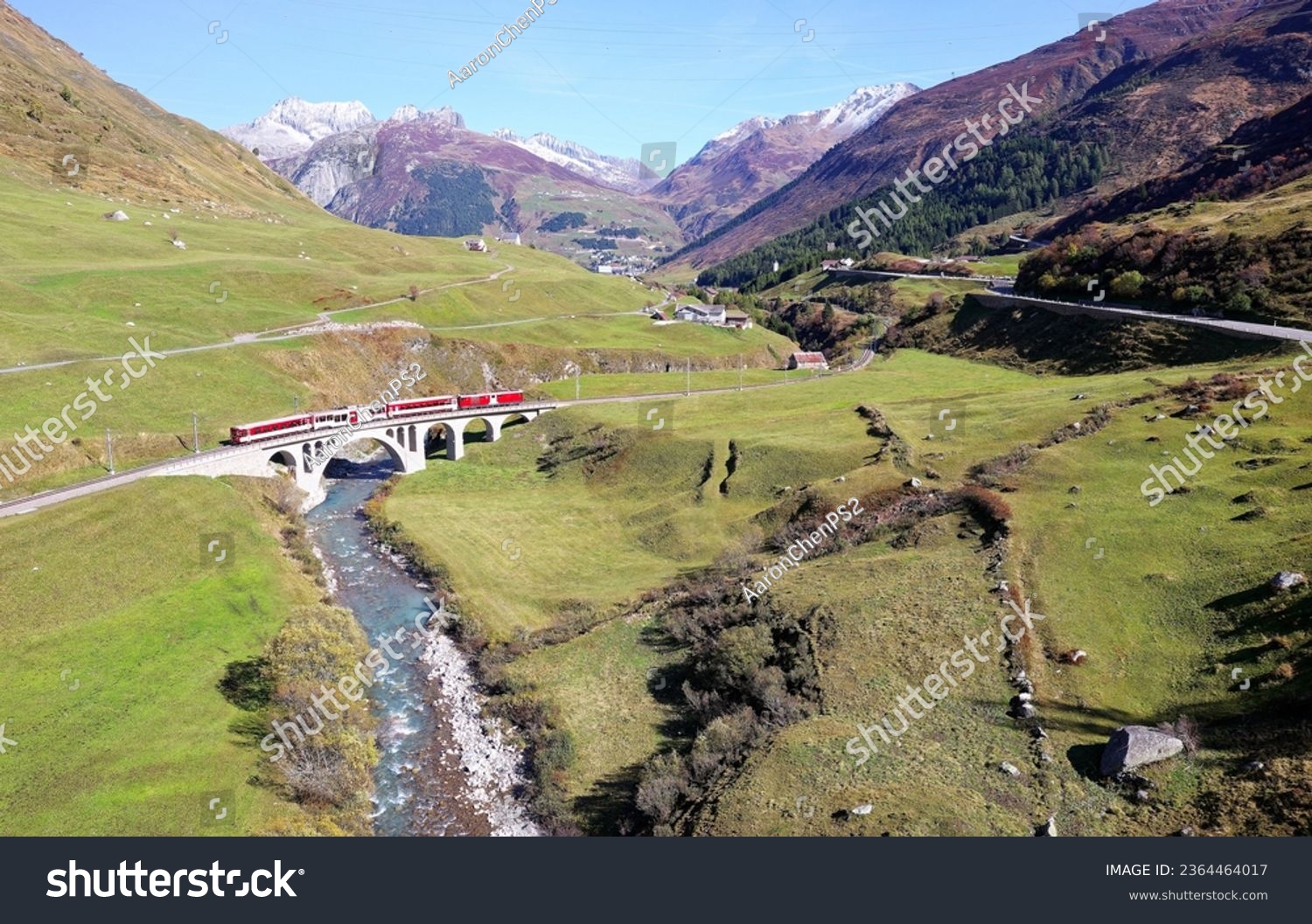 A train of Matterhorn Gotthard Bahn travels across a viaduct over a stream in the green valley between alpine mountains on a beautiful sunny day, in Hospental village near Andermatt, Uri, Switzerland #2364464017