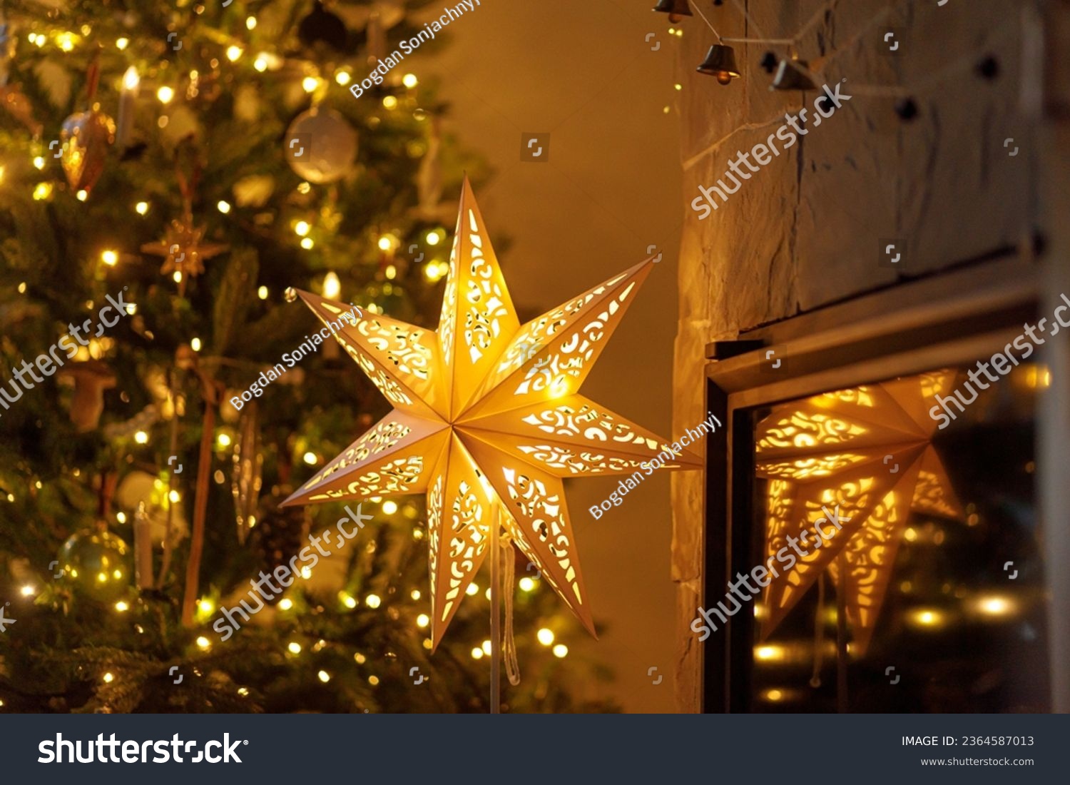 Atmospheric christmas eve. Stylish christmas illuminated star, decorated christmas tree with golden lights and festive decor on fireplace mantel in scandinavian room. Merry Christmas! #2364587013