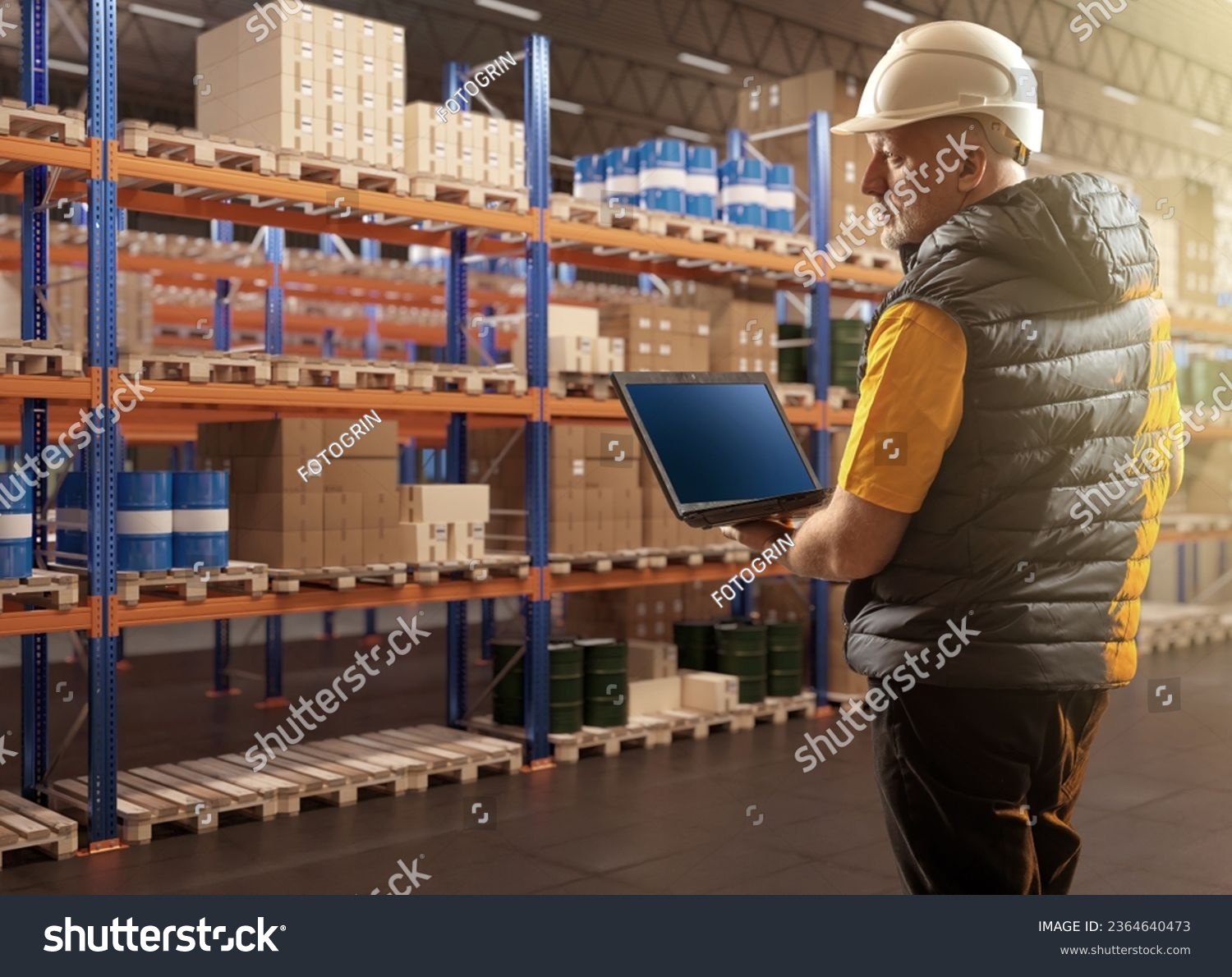 Man in warehouse. Logistics center employee. Worker with laptop in warehouse. Man inspects warehouse racks. Barrels and boxes are stored on pallets. Businessman with laptop in storage building #2364640473