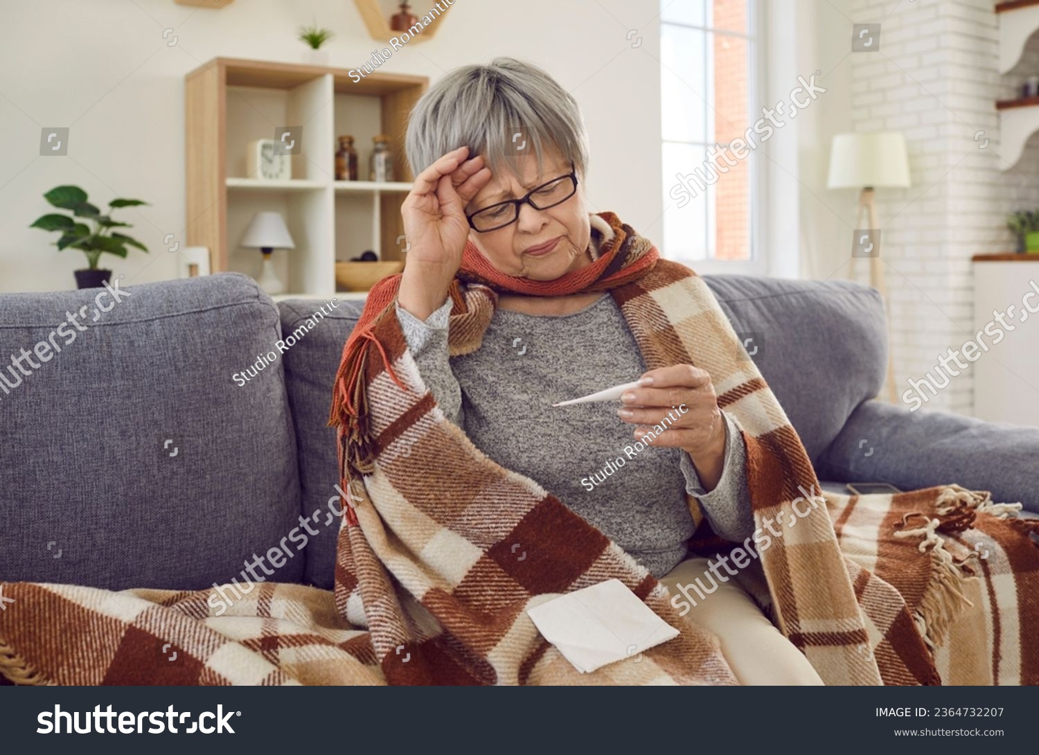 Sick senior woman checking her temperature wrapped in a blanket on the sofa suffering from headache. Elderly person with seasonal flu or cold feel unhealthy with influenza at home. #2364732207