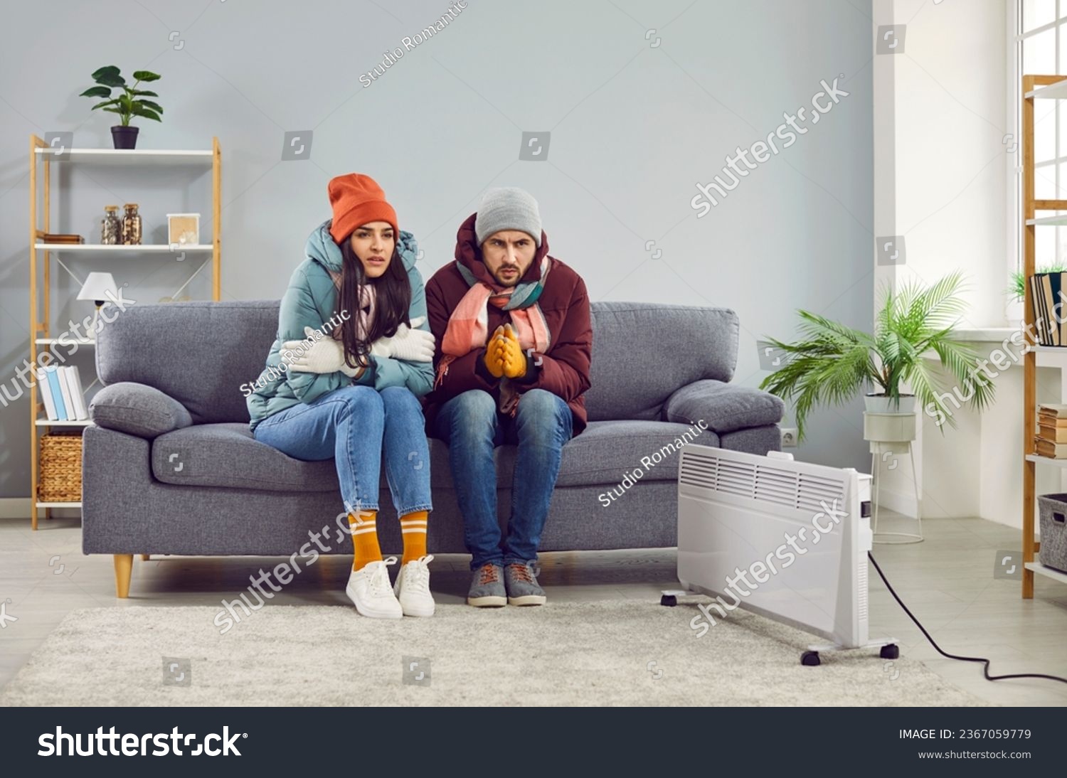 Freezing people in winter jackets sitting on sofa at home. Sad warmly closed husband abd wife feeling cold and trying to warm up. Heating problems, power crisis concept #2367059779
