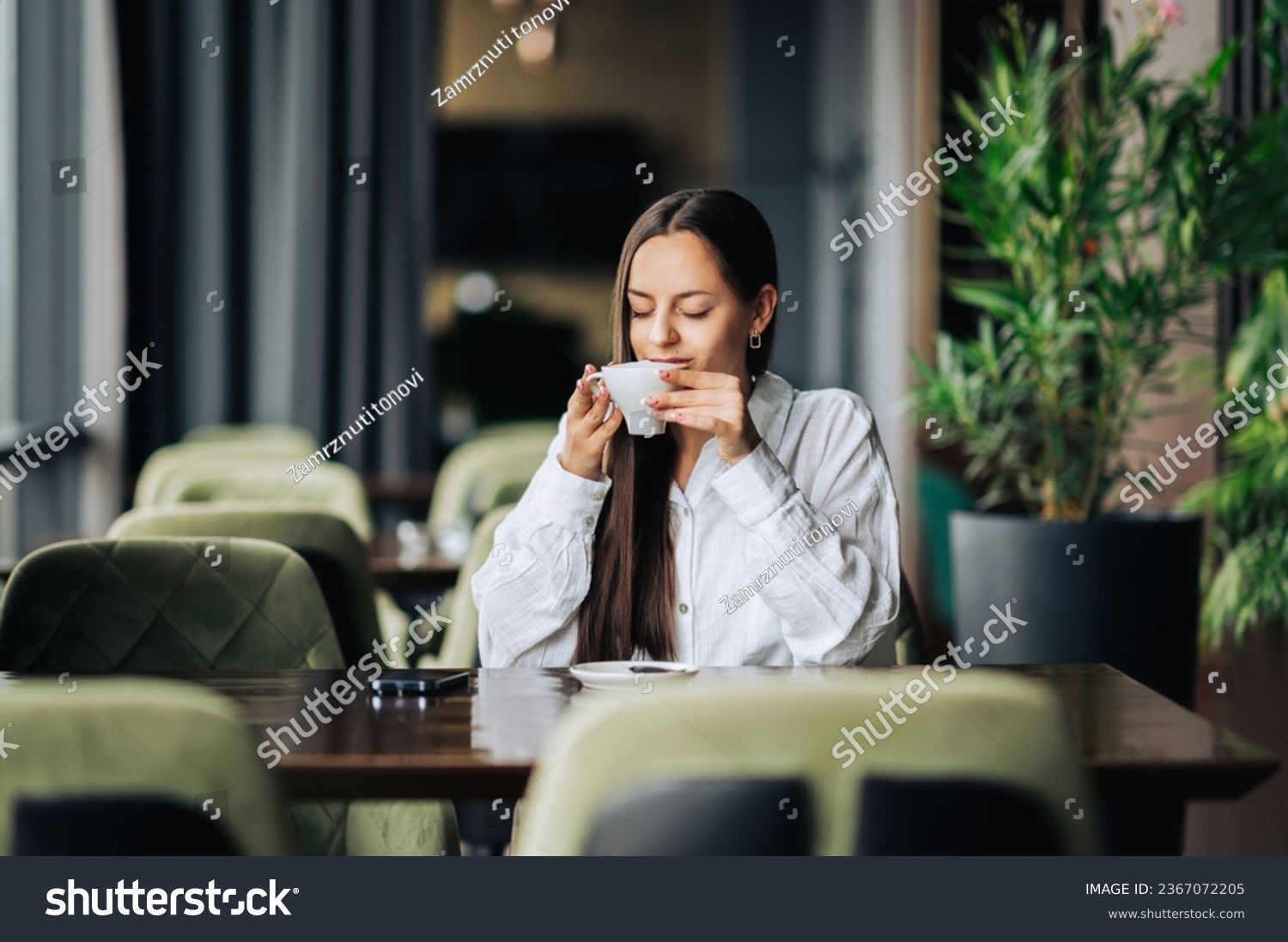 A cheerful brunette is sitting in a cafe and sipping fresh espresso and enjoying morning moments. A young trendy woman is enjoying her coffee in a cafeteria. #2367072205