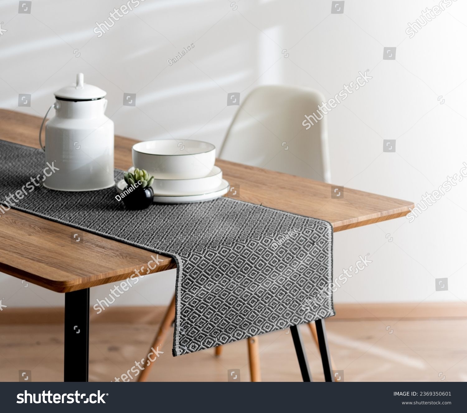 Modern Dining Room Featuring a Wooden Dining Table Adorned with a Grey Casual Linen Textured Table Runner, Teapot, and White Plates beside a White Plastic Dining Chairs, White Wall, Close Up. #2369350601