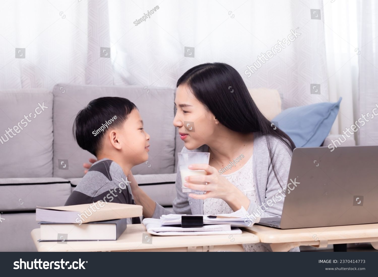 Side view Asian boy kid give mom glass of milk with love tenderness care, son bring cup of milk to mother and sit together while female working on technology laptop in living room happy mom lifestyle #2370414773