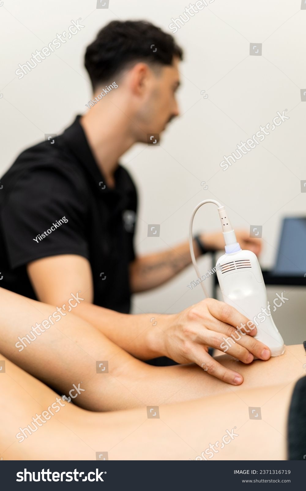 A male physical therapist is performing an ultrasound on a patient's femoral to detect pain. The probe in the client's leg is in the foreground. Concept of ultrasound in physiotherapy clinics. #2371316719
