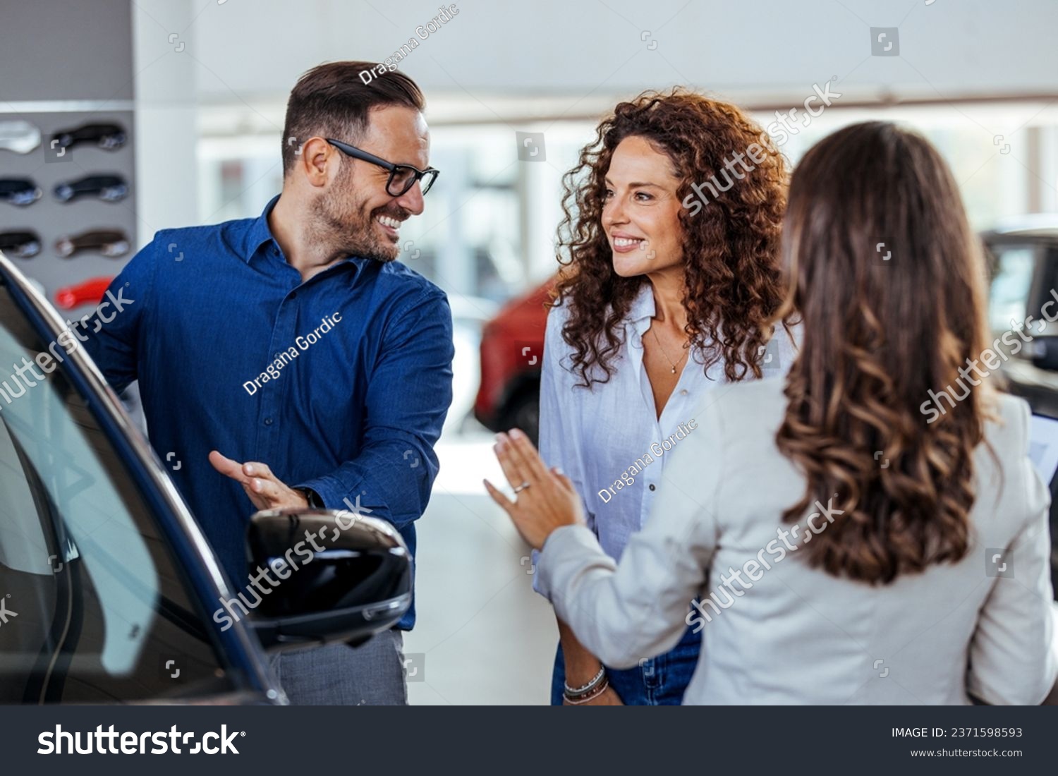 A car salesman is showing new cars to a couple. Car sales business. Manager talking to couple, showing them new auto at dealership shop. Young family selecting vehicle, looking at modern automobile  #2371598593