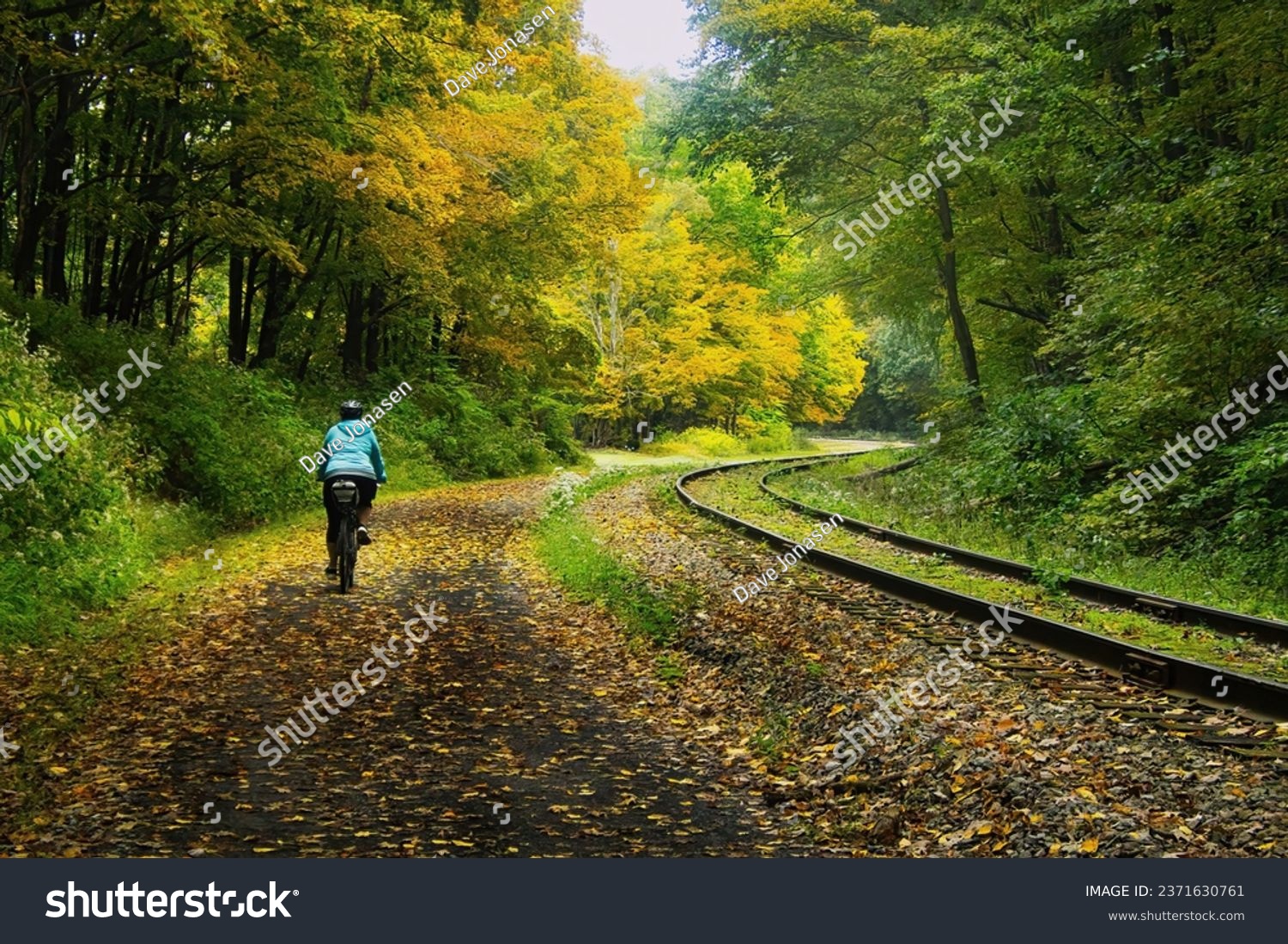 Landscape of a cyclist, seen from behind, on the Great Allegheny Passage Trail curving past railroad tracks and a forest on a moody early Autumn day. #2371630761