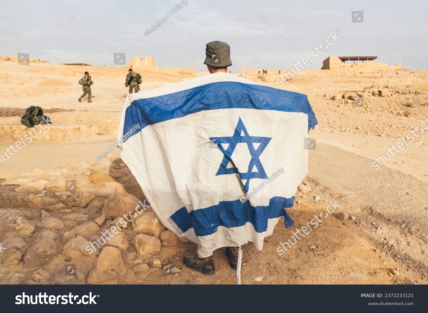 Back view of Israeli soldier with white and blue flag of Israel against the background of the desert and rocks.. Military men protecting promissed land. National flag of Israel. War conflict #2372233121