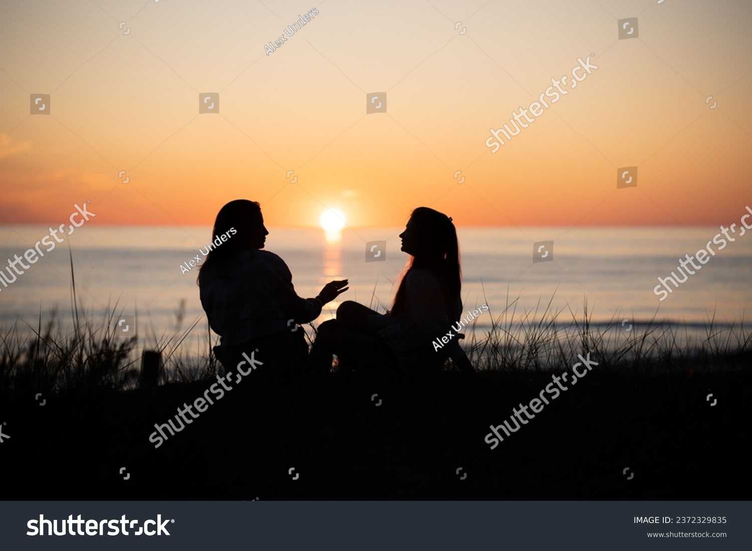 Silhouette of two female friends enjoying a conversation while sitting and watching the sunset on a sea beach. Back view #2372329835