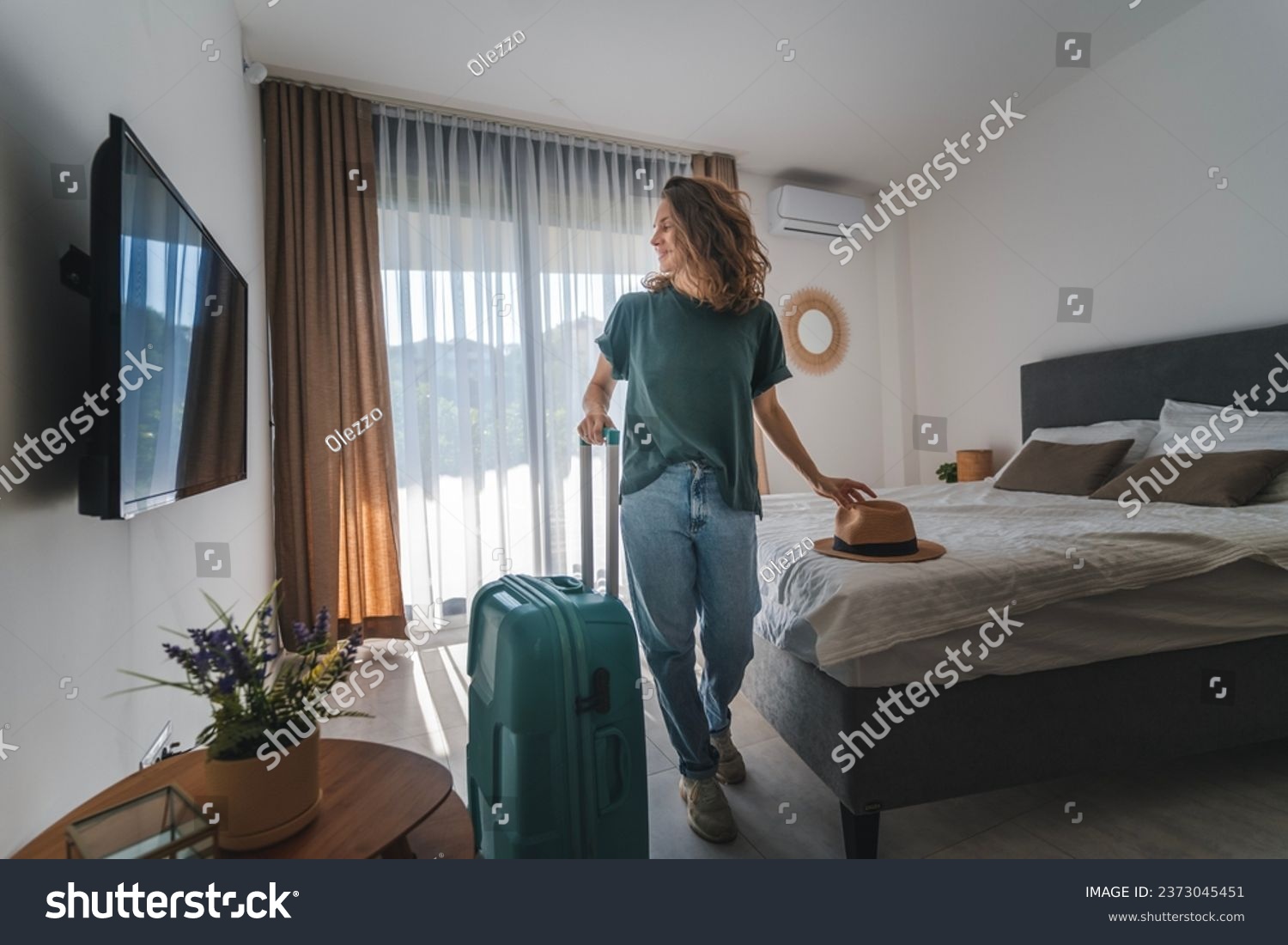 A young woman in a hat with a suitcase checks into a hotel room. Booking and Accommodation travel  #2373045451