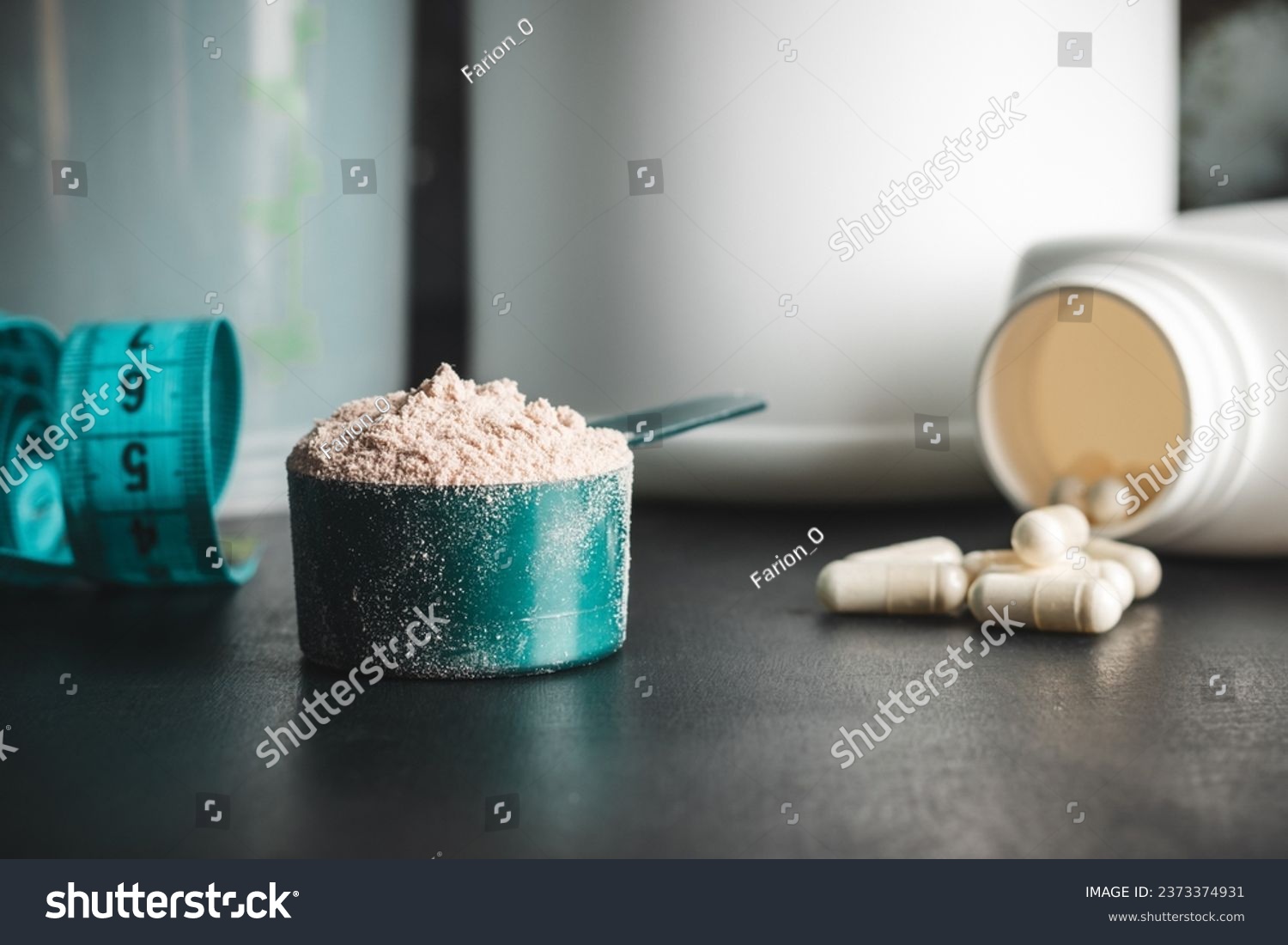 Chocolate whey protein powder in measuring spoon, white capsules of amino acids, vitamins and creatine, measuring tape, plastic shaker on dark background. bodybuilding food supplements. #2373374931