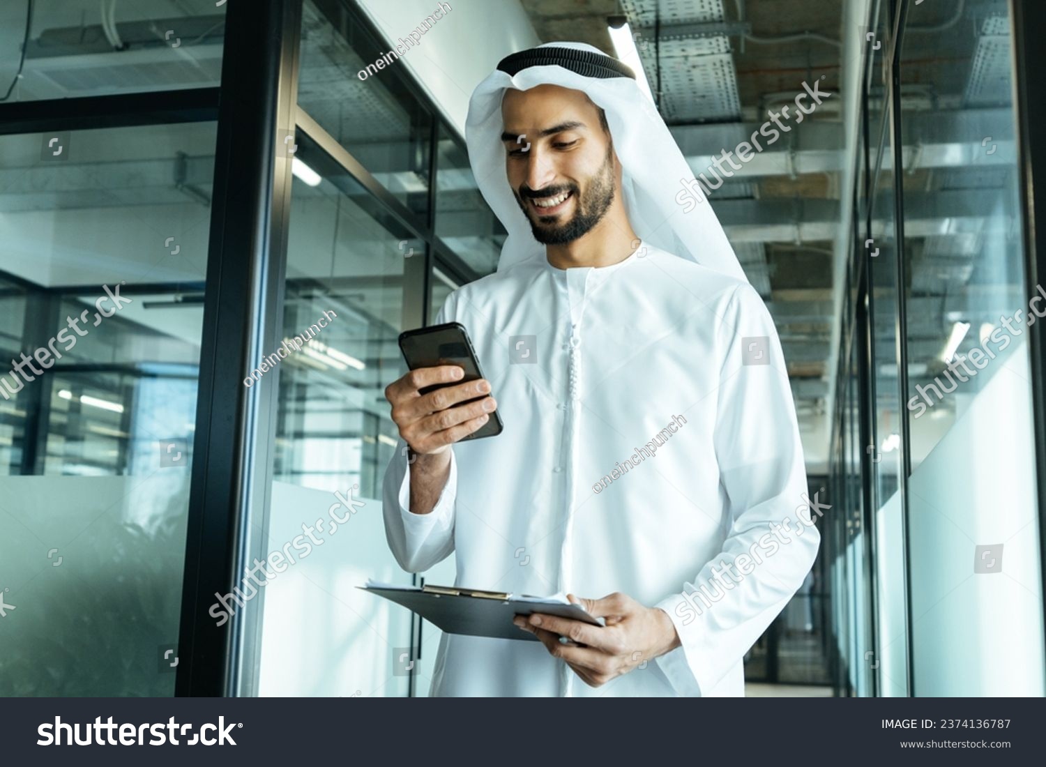 handsome man with dish dasha working in his business office of Dubai. Portraits of a successful businessman in traditional emirates white dress. Concept about middle eastern cultures. #2374136787
