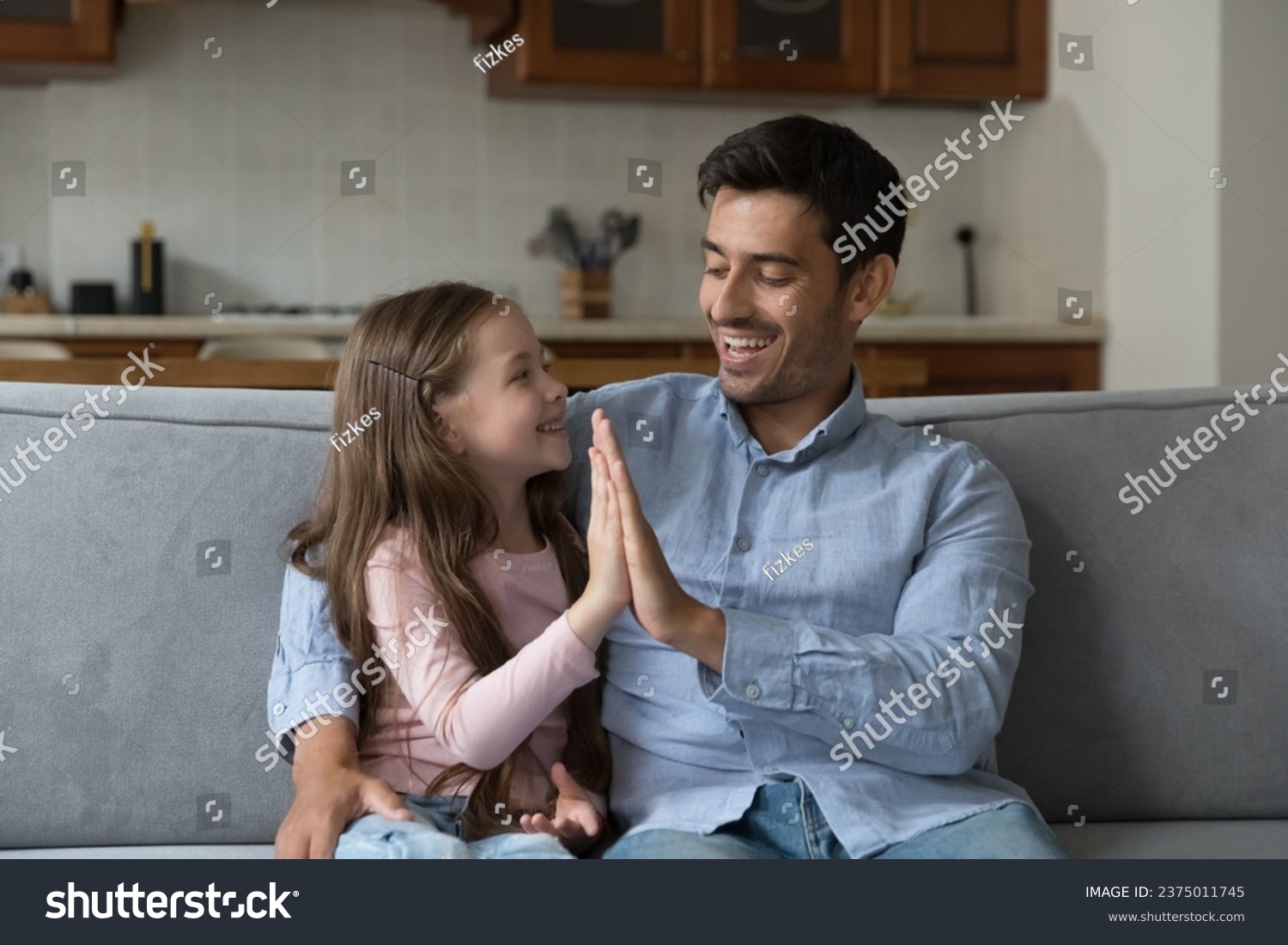 Proud kid girl and happy father giving high five on home couch, hugging, talking, laughing, enjoying leisure. Father giving praise, support to little daughter. Family, parenthood, fatherhood concept #2375011745