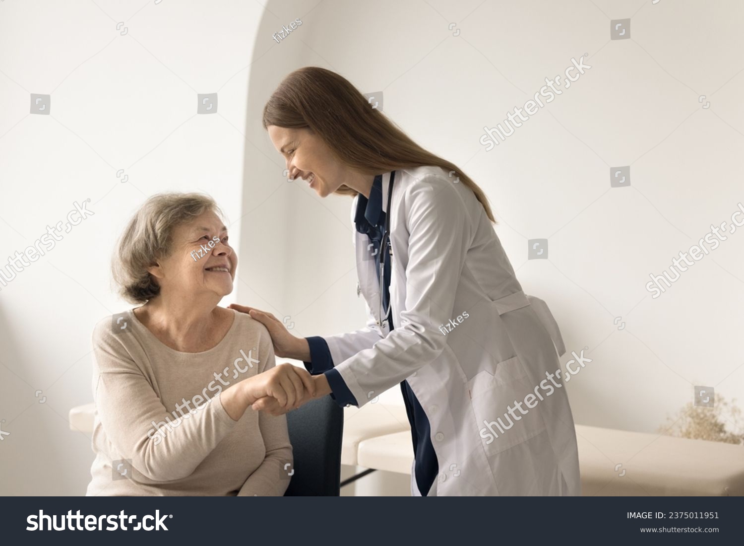Smiling retired woman visiting doctor in hospital, get good news after medical check-up. Young female general practitioner holds patient hand, touch shoulder, encourages, give psychological support #2375011951
