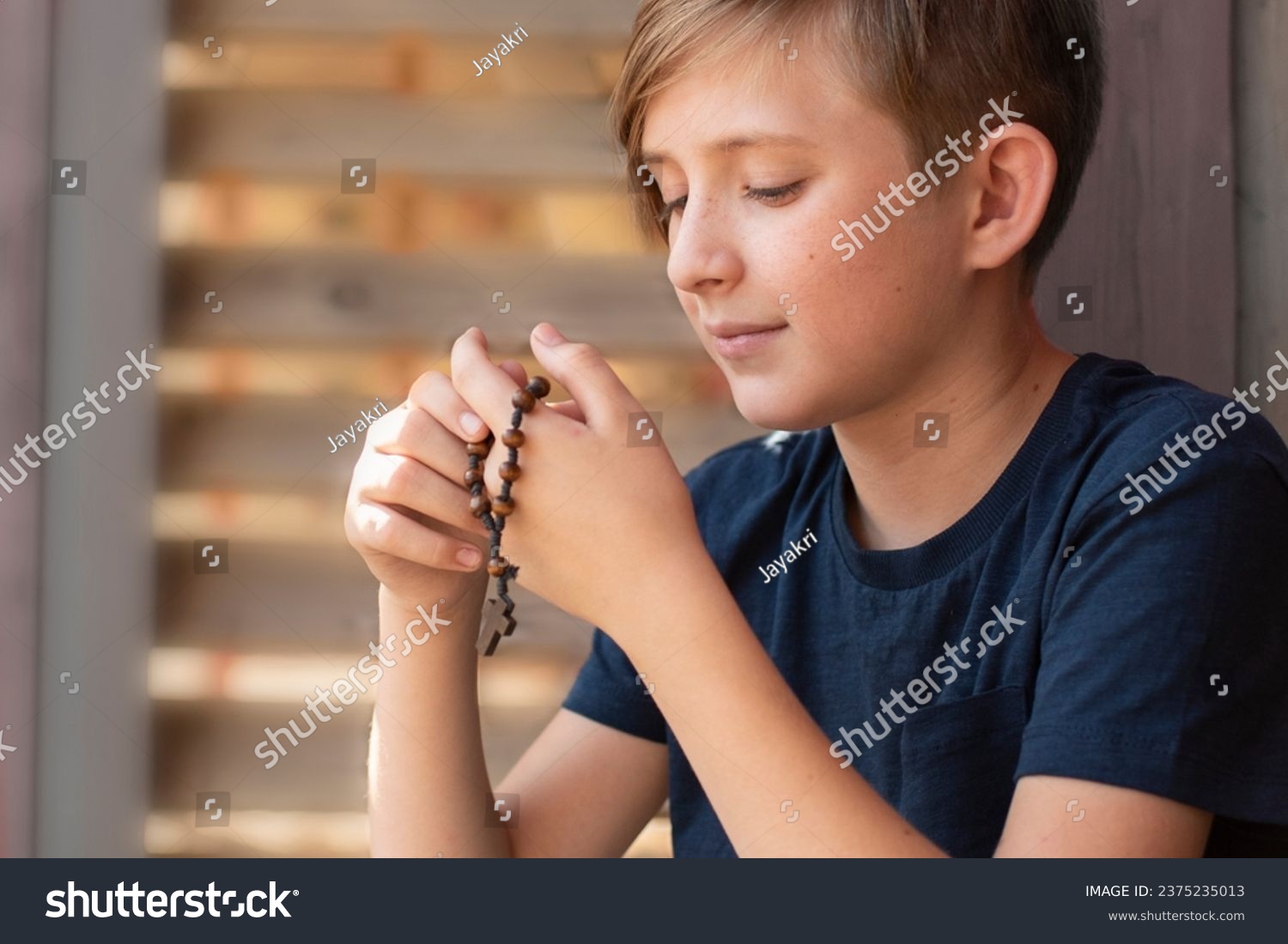 An 11 year old Catholic boy reads the rosary prayer, holds a wooden rosary with 10 beads in his hands. portrait of a boy with a wooden Catholic rosary during prayer. #2375235013
