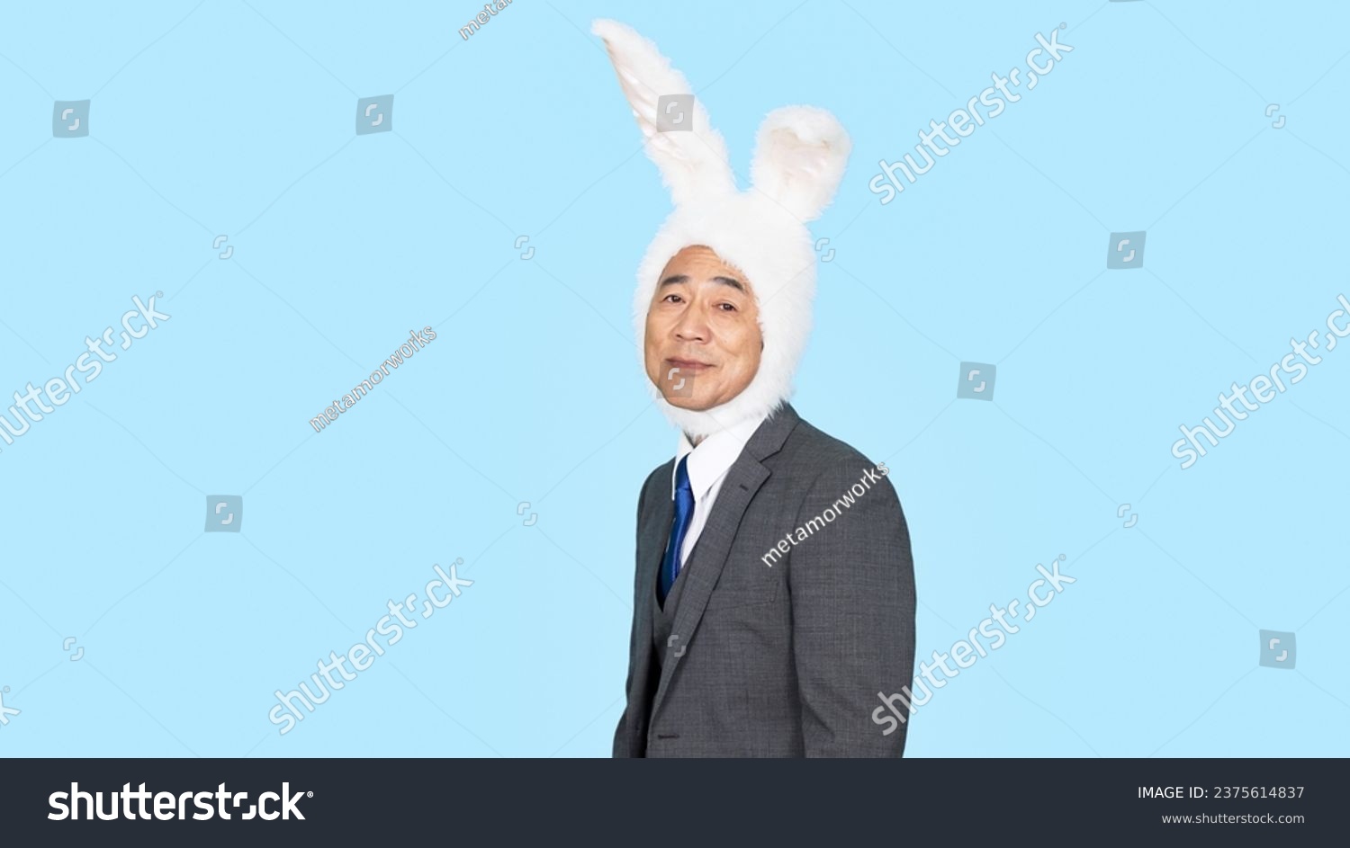 A middle-aged Asian man in a business suit wearing a rabbit headgear in front of a blue background. Party entertainment. Halloween party. #2375614837