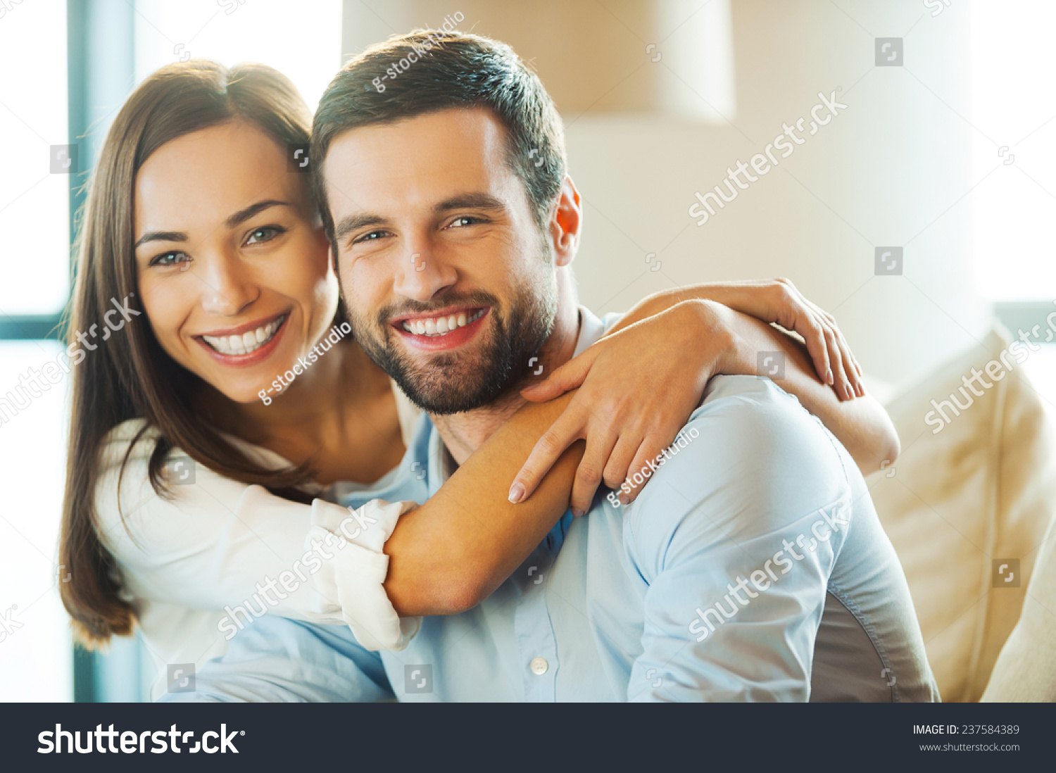 Enjoying every minute together. Beautiful young loving couple sitting together on the couch while woman embracing her boyfriend and smiling  #237584389