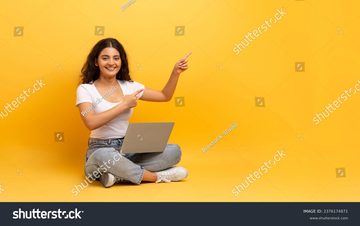 Remote job opportunities, freelance. Happy beautiful curly young indian woman student wearing casual outfit sitting on floor with laptop on her lap, pointing at copy space, yellow background, panorama #2376174871