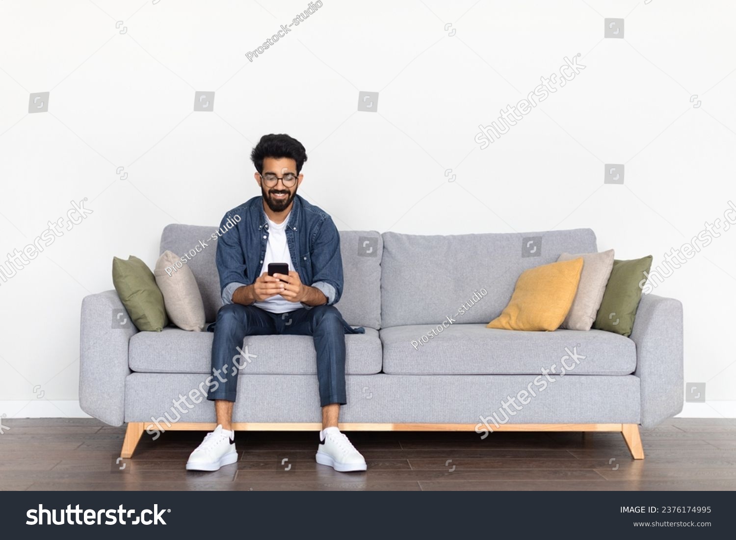 Casual Indian guy, wearing a casual outfit, sits on a cozy sofa, smiling while exploring phone, against a pristine white backdrop, illustrating a moment of joyful digital communication, copy space #2376174995