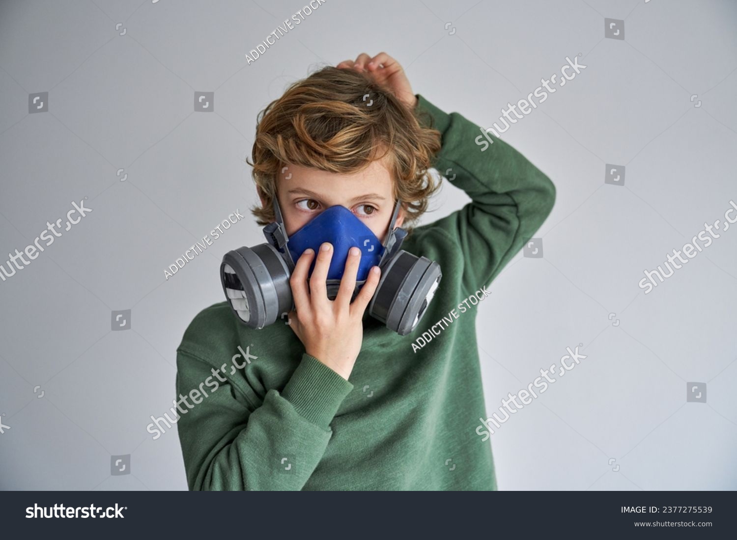 Thoughtful little boy with blond hair in casual clothes putting on protective respirator and looking away against gray background #2377275539