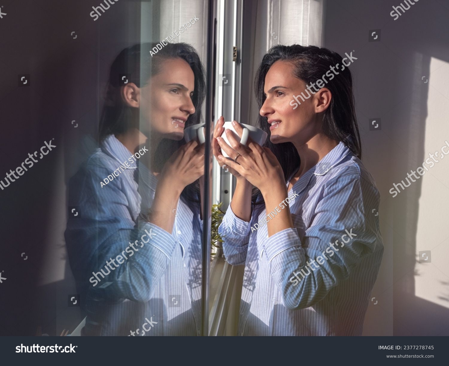 Side view of charming female with long dark hair and in pajamas drinking hot coffee from cup while standing near window in sunny morning #2377278745
