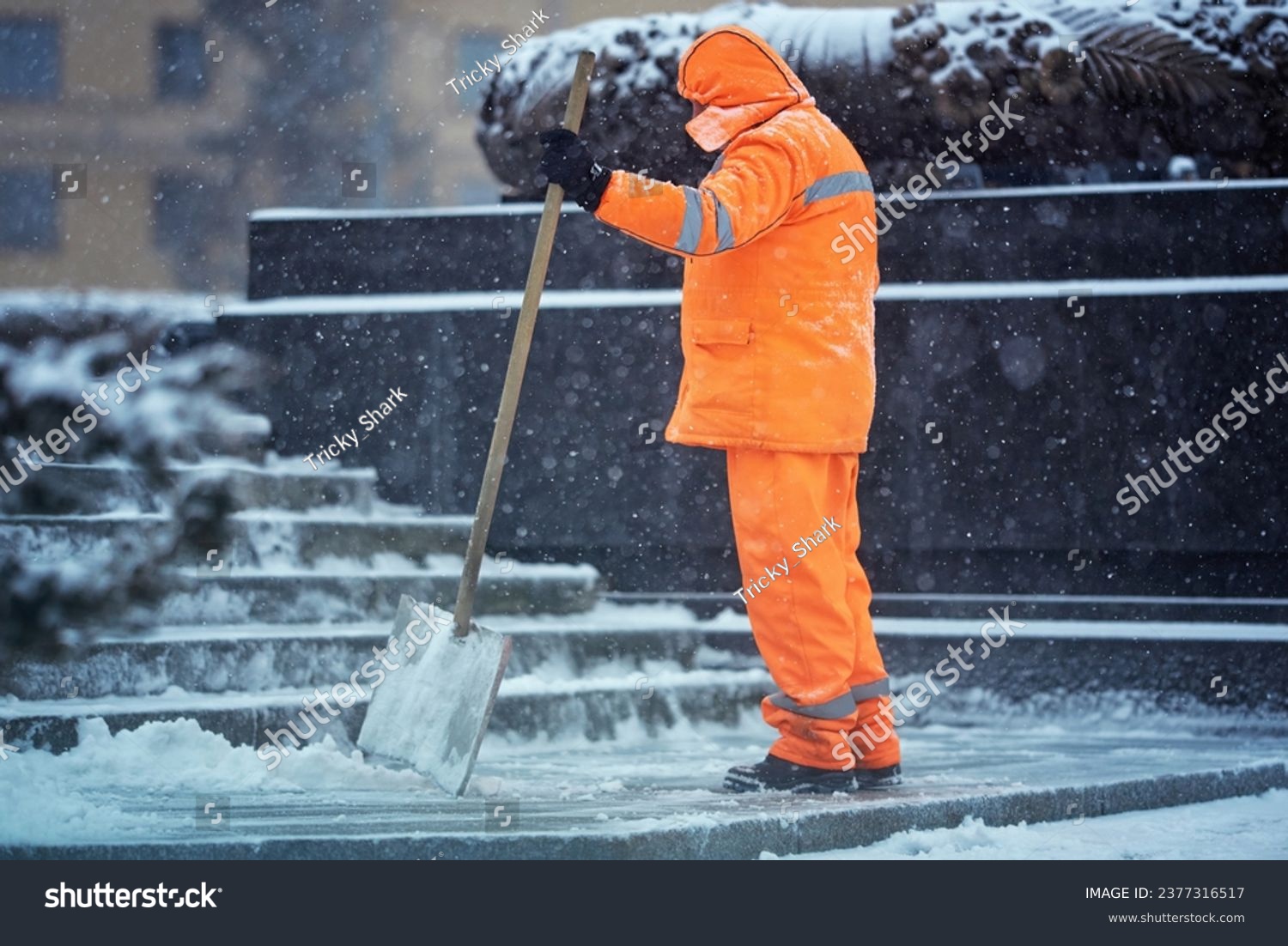 Utility man with snow shovel clear snow during blizard, snow removal work. Municipal worker in uniform with snowshovel in hands clean snowy sidewalk and walkway during winter storm #2377316517