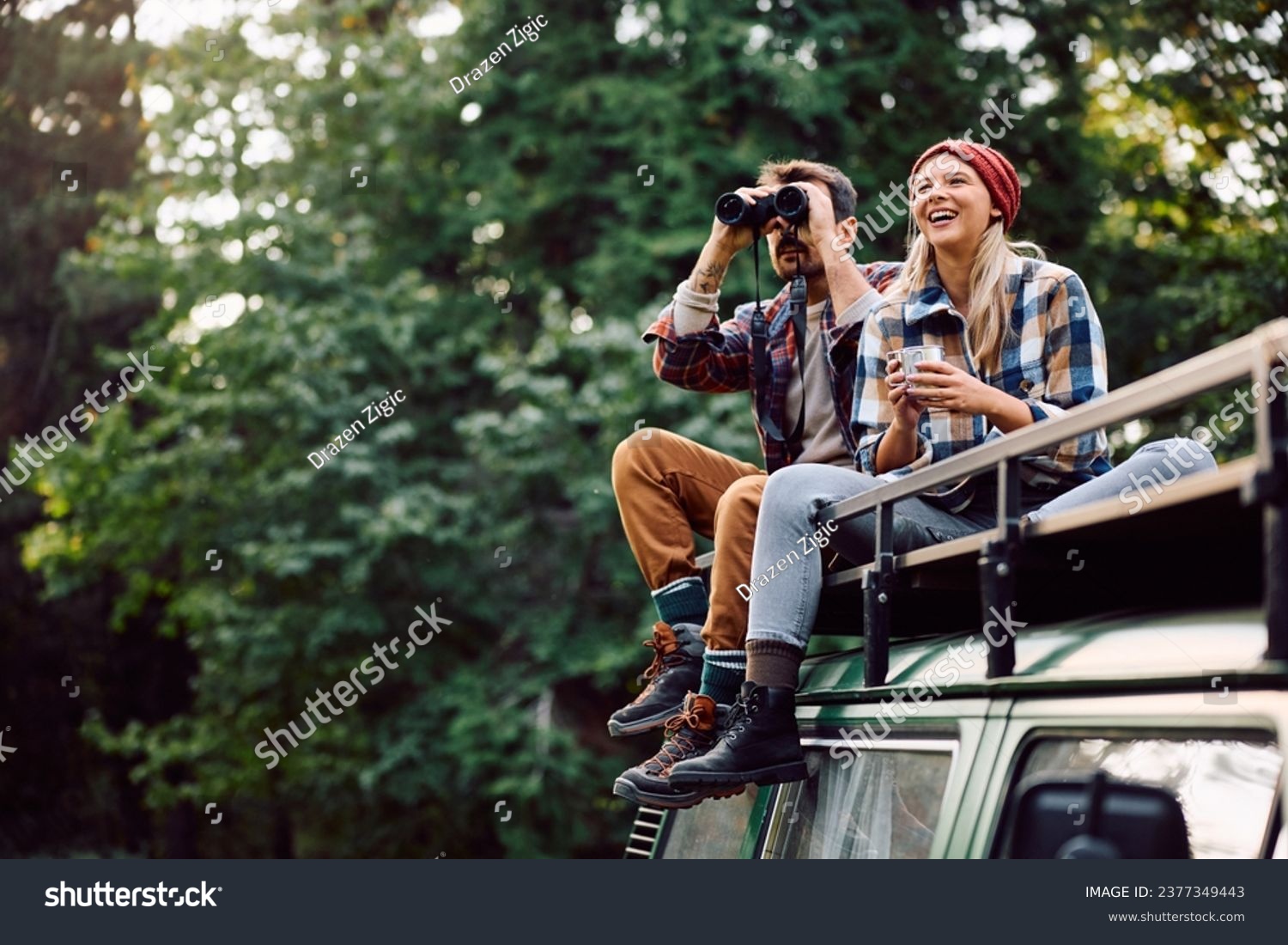 Happy woman and her boyfriend using binoculars while sitting on roof of their camper trailer and spending a day in the woods. Copy space. #2377349443