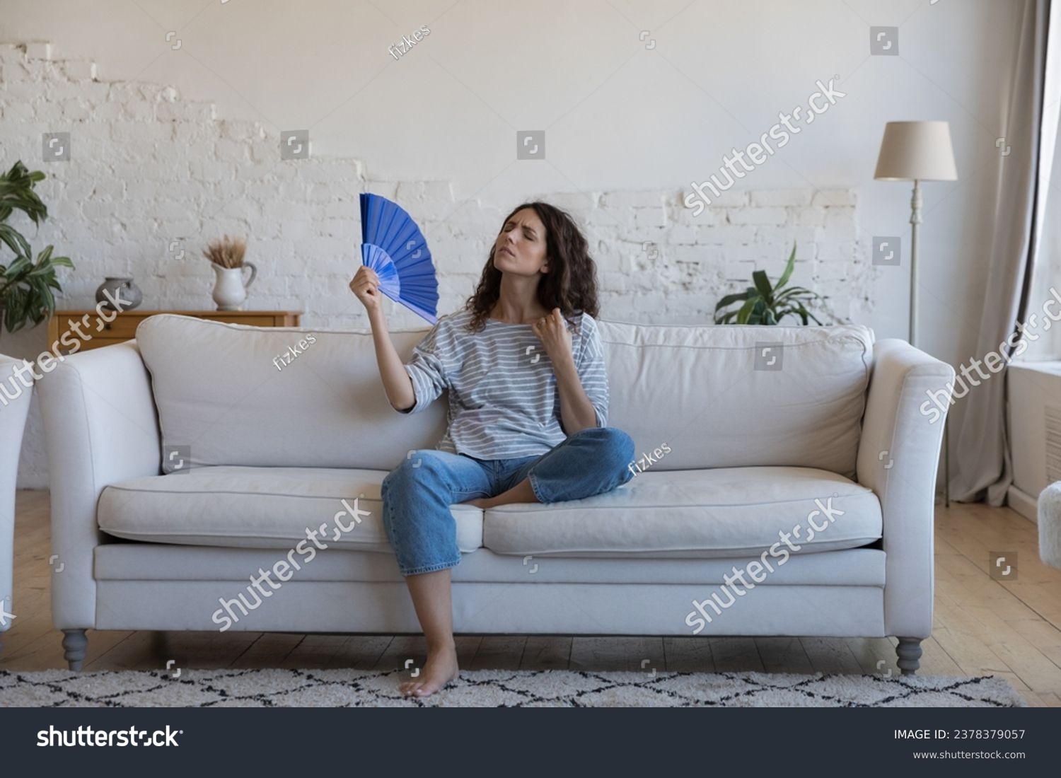 Exhausted frustrated Hispanic woman waving paper handheld fan, cooling hot air, suffering from heat attack, headache, hypoxia, stuffy air, sitting on home couch in living room #2378379057