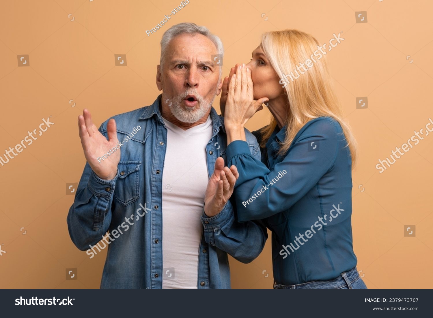 Attractive mature blonde woman sharing secret or whispering gossips into her husband ear, beige studio background. Loving elderly spouses sharing rumors. Communication in marriage #2379473707