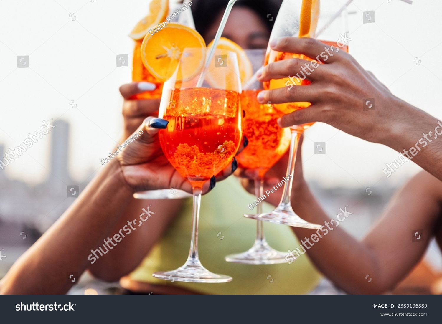 Close up of female and male hands holding elegant glasses with long stem of aperol and clink them. Making a celebratory toast with delicious citrus alcohol cocktails with slice of orange. Party. #2380106889