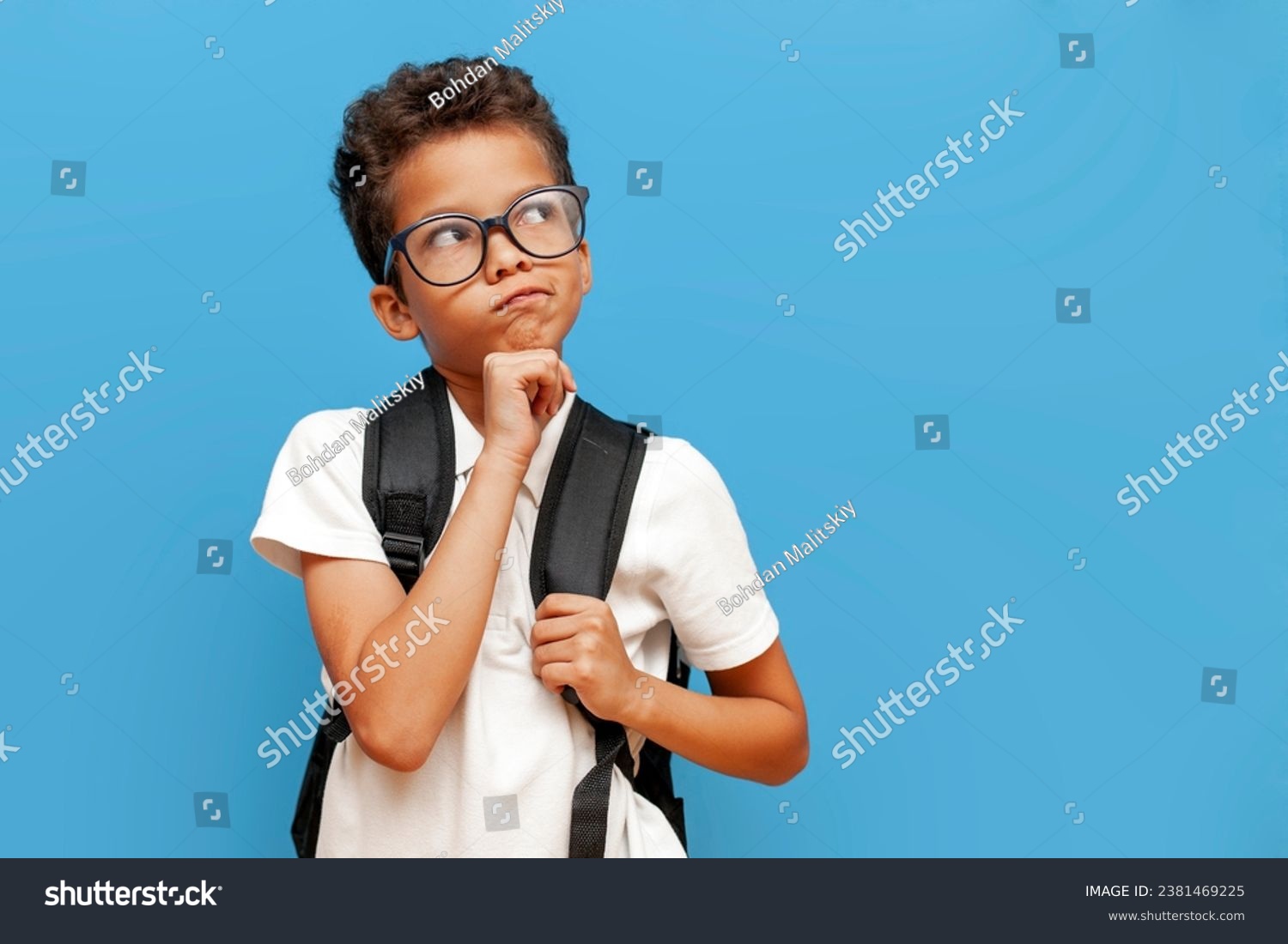 pensive african american teenager boy in glasses with backpack thinks and plans on blue isolated background, curly schoolboy imagines daydreaming and looks away #2381469225