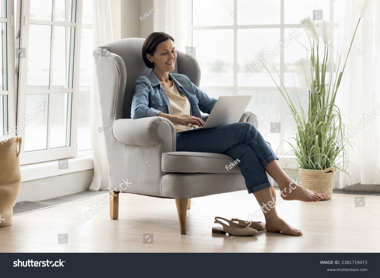 Middle-aged woman relax on comfortable armchair in living room with laptop, watch movie on digital streaming on-line services, spend time on internet, make order, enjoy leisure and modern tech usage #2381719473