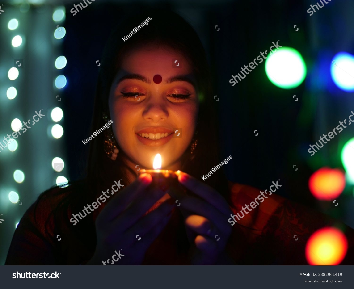 Pretty young female holding an oil-lit lamp in a dark room - festival of Diwali celebration . Smiling Indian woman with a Diwali lamp in hand - an auspicious day, Diwali preparation, bokeh shot #2382961419