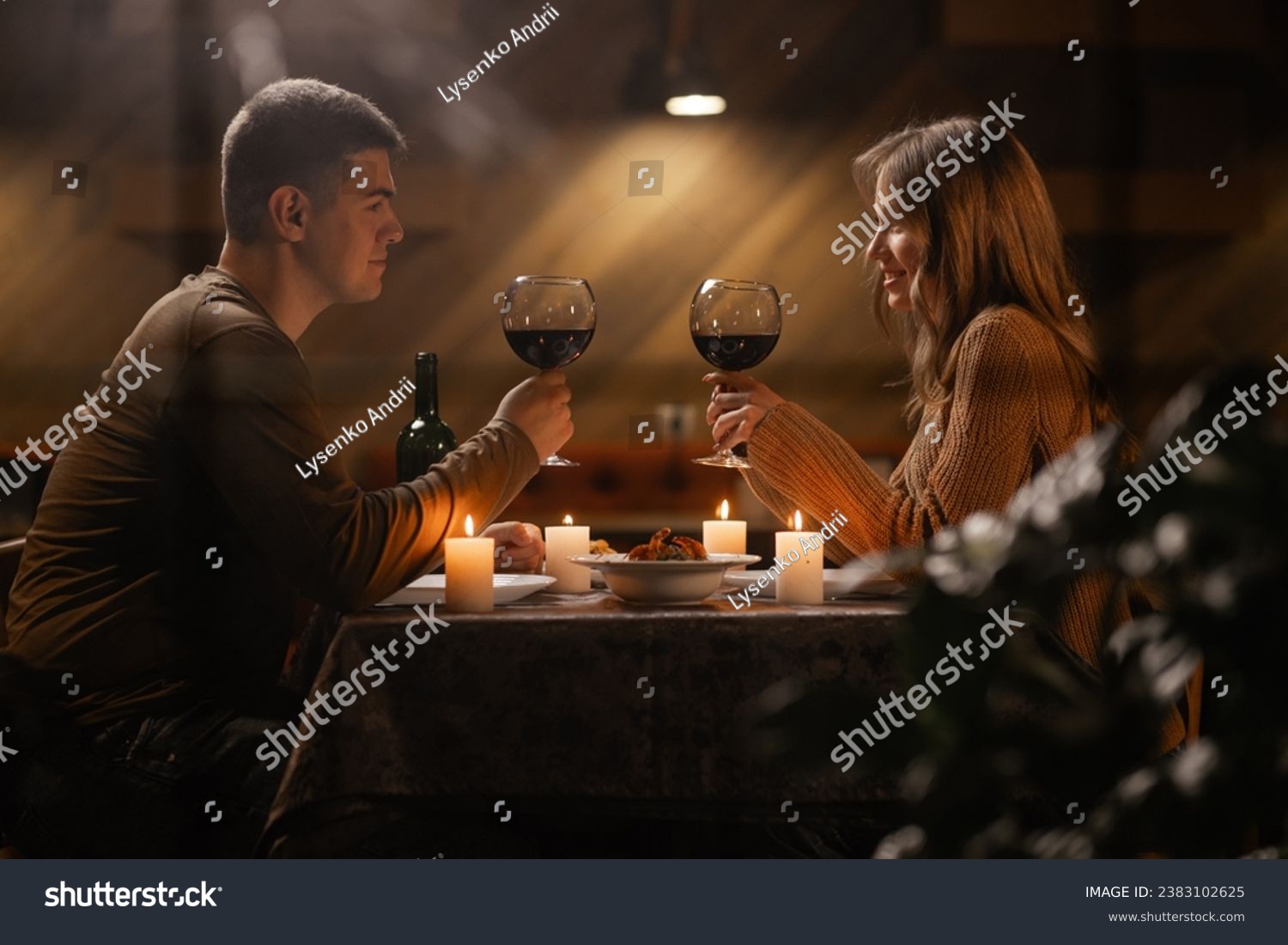 Trough window view on young couple in love drinking wine, celebrating Valentines day dining at home, having romantic dinner date with candles. Copy space #2383102625