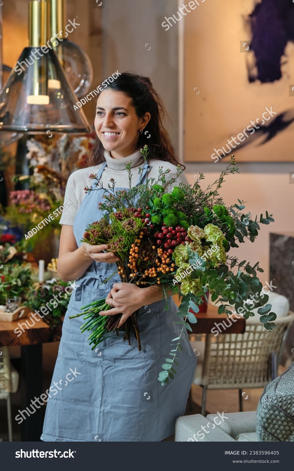 Smiling young woman in blue apron touching arranged flower bouquet and looking away while working in floral shop on blurred background #2383596405