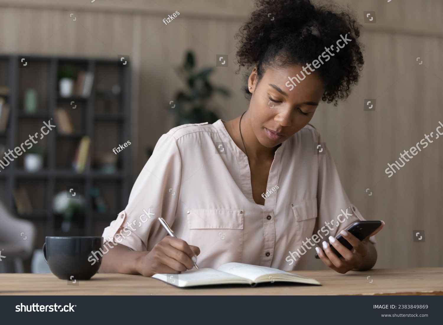 Serious African American millennial student watching learning webinar, virtual training, online workshop on smartphone, writing notes in notebook, studying from home, taking remote course #2383849869