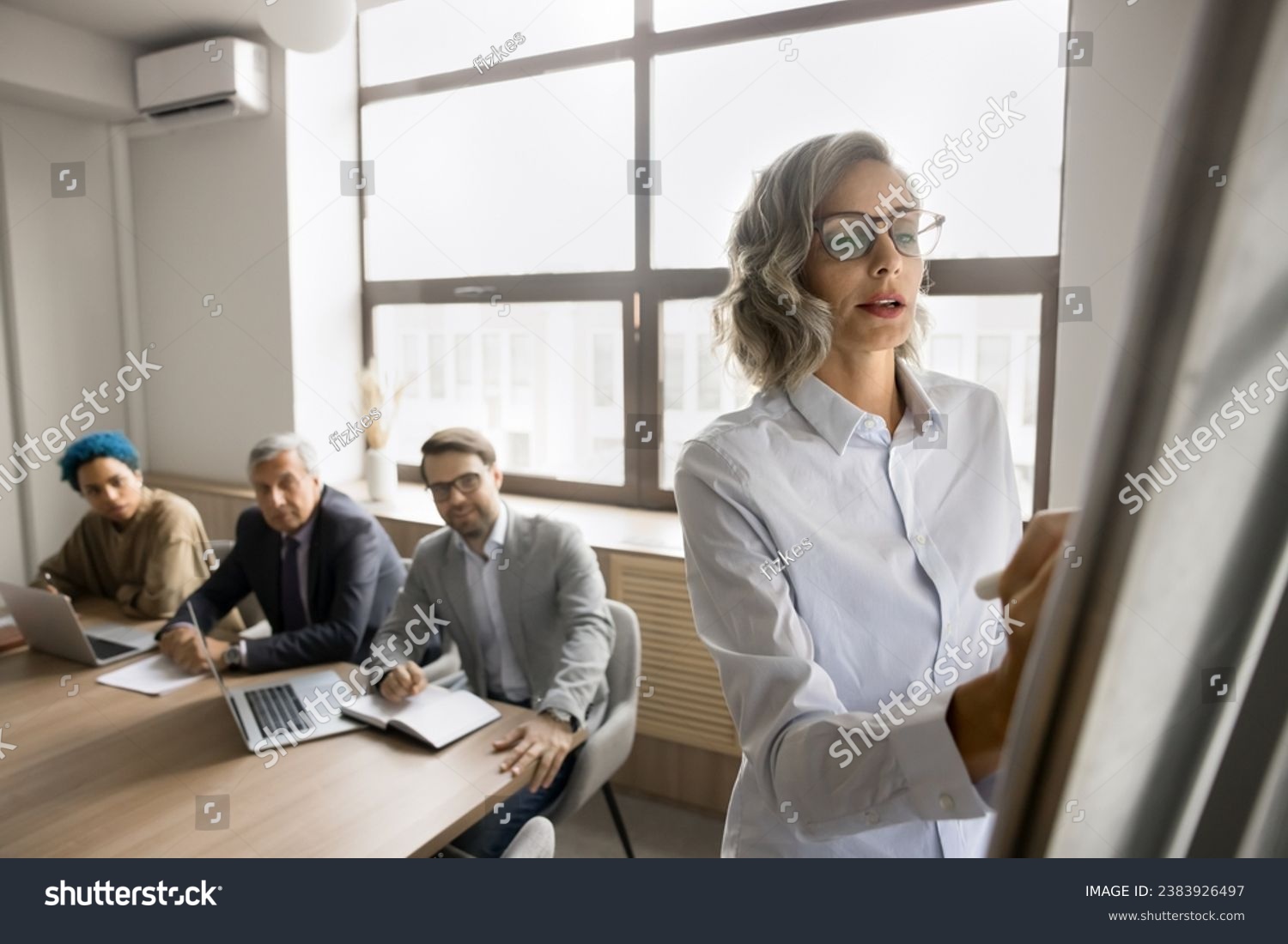 Serious pretty mature female business leader writing on whiteboard on meeting with multiethnic team, explaining project plan, strategy, sharing ideas with office colleagues, giving report, seminar #2383926497