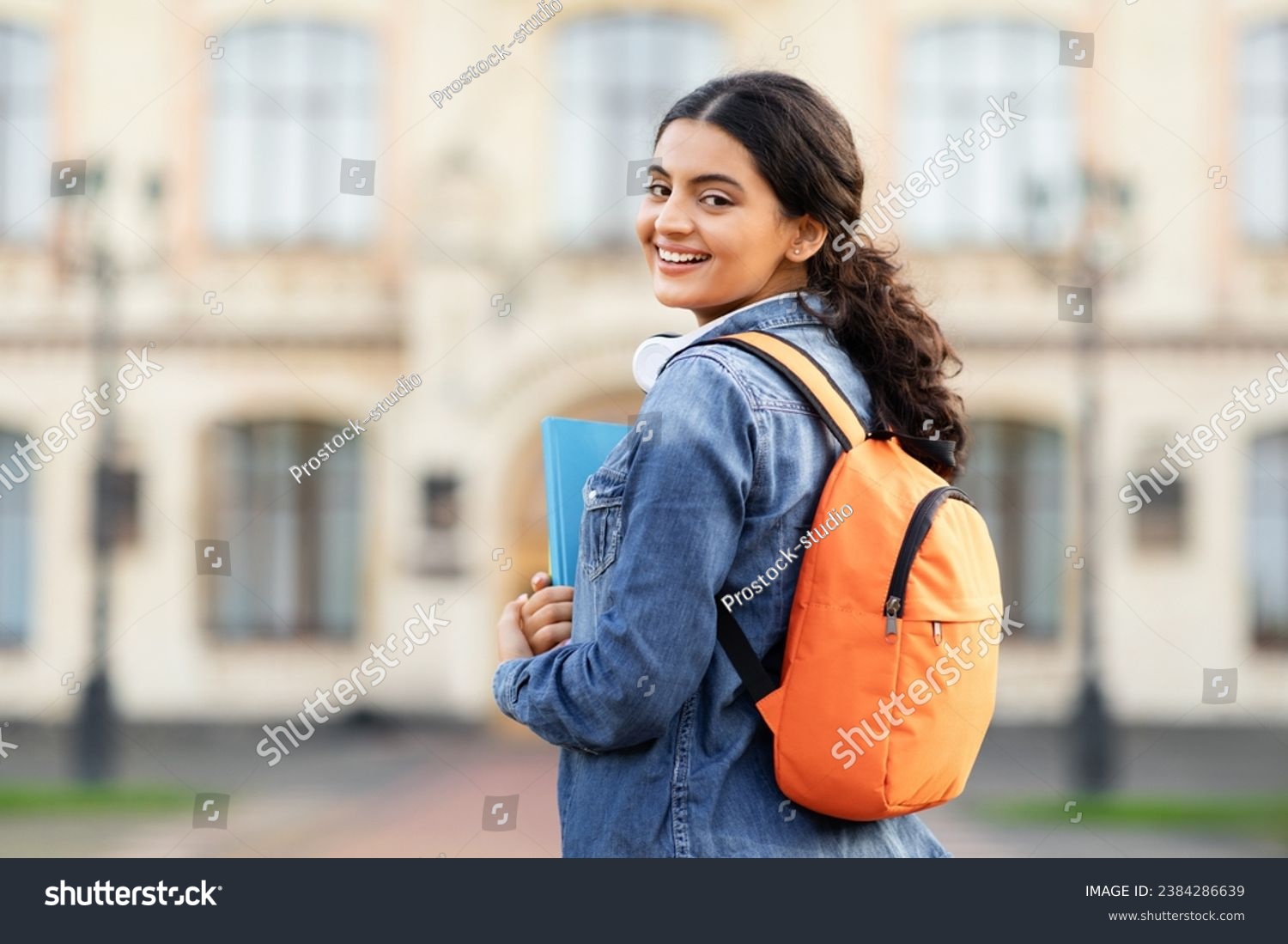 A radiant young Indian scholar with her academic books and bag, posing outside her educational institution, offering a warm smile to the lens. Space for caption, emphasizing education. #2384286639
