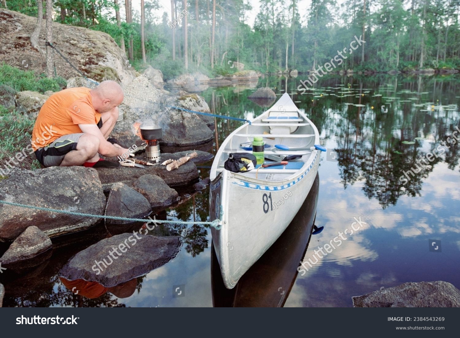 A man prepares breakfast on a small camping stove next to a metal canoe by a lake in Sweden #2384543269