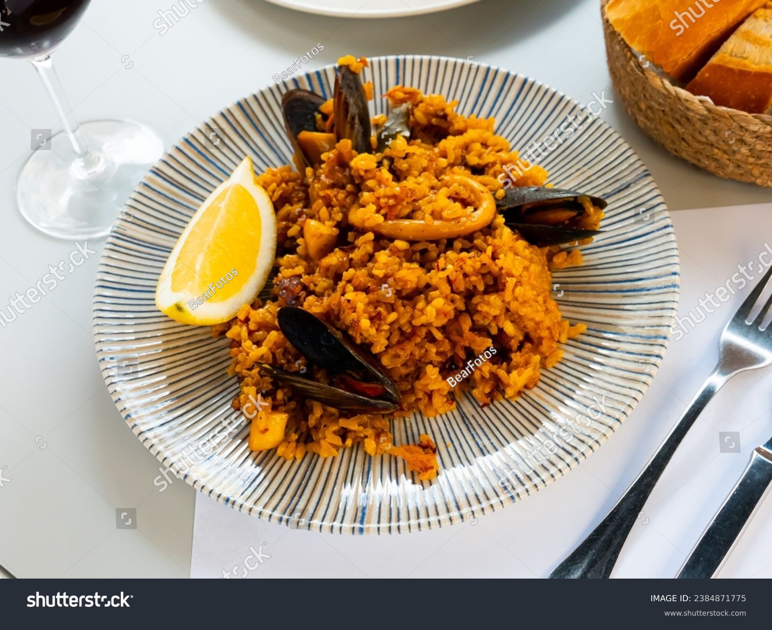 Appetizing racy seafood paella with mussels and squid rings served with lemon slice and glass of red wine. Traditional Spanish cuisine. #2384871775