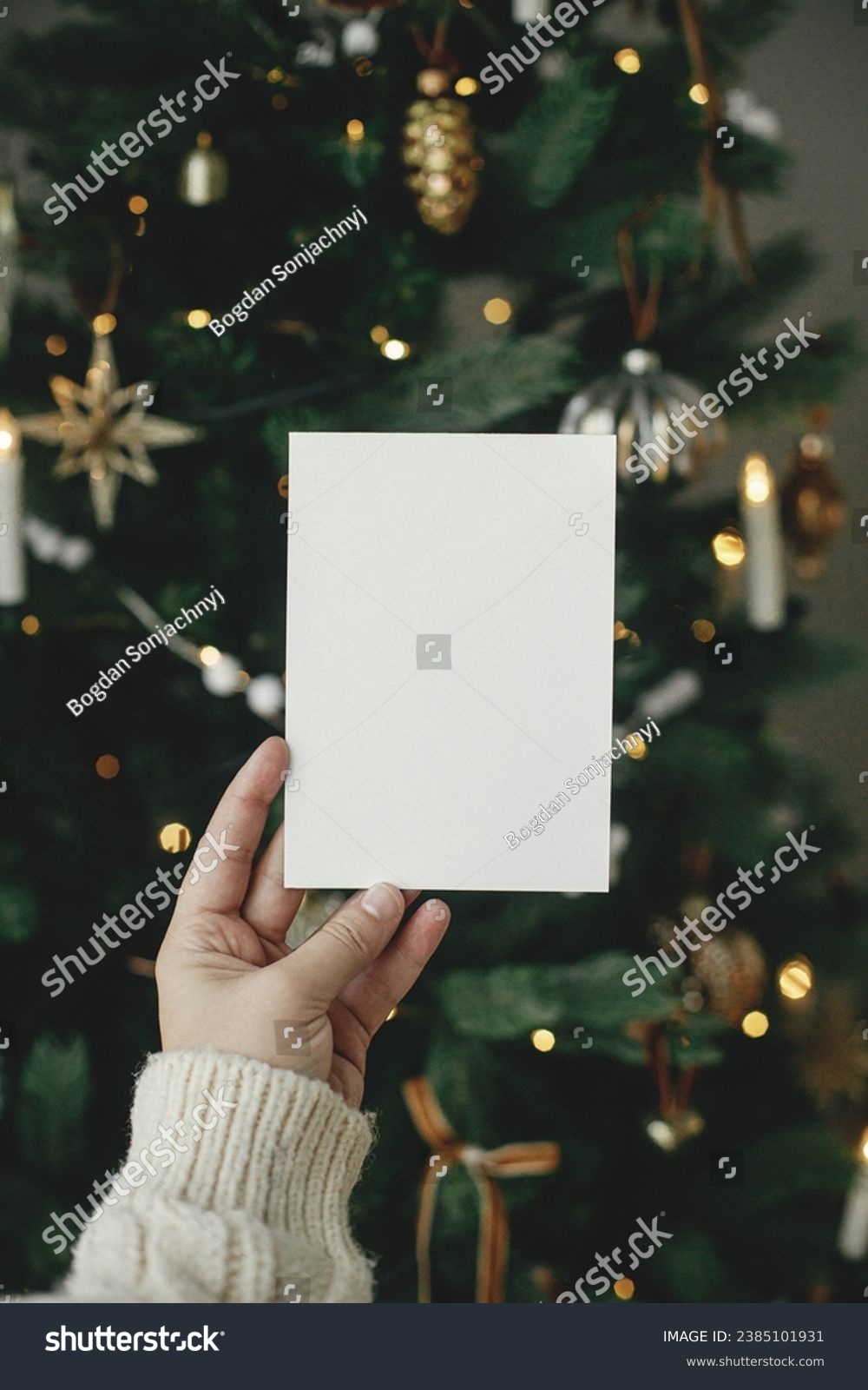 Christmas card mock up. Hand holding empty greeting card on background of stylish decorated christmas tree with golden lights. Space for text. Season greetings template and vintage ornaments #2385101931