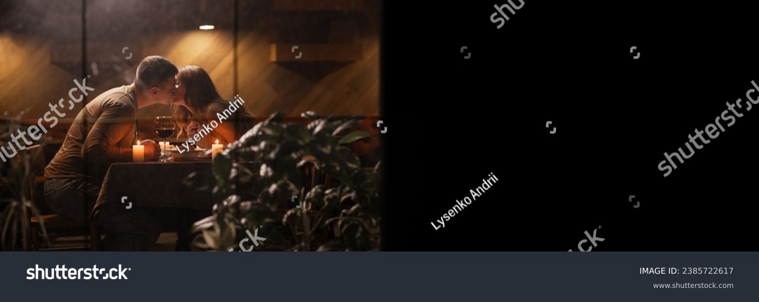 Romantic young couple kiss while during dinner at dining table celebrating Valentine's Day, view through window. Banner. Copy space #2385722617