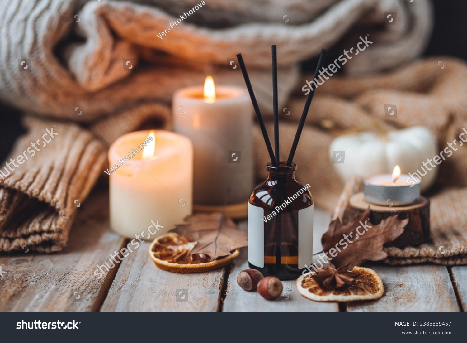 Hello autumn, cozy slow living. Pumpkin, aromatic candle, warm sweaters, dry fall leaves, cinnamon, anise in golden lights with bokeh on white wooden background. Thanksgiving apartment decor #2385859457